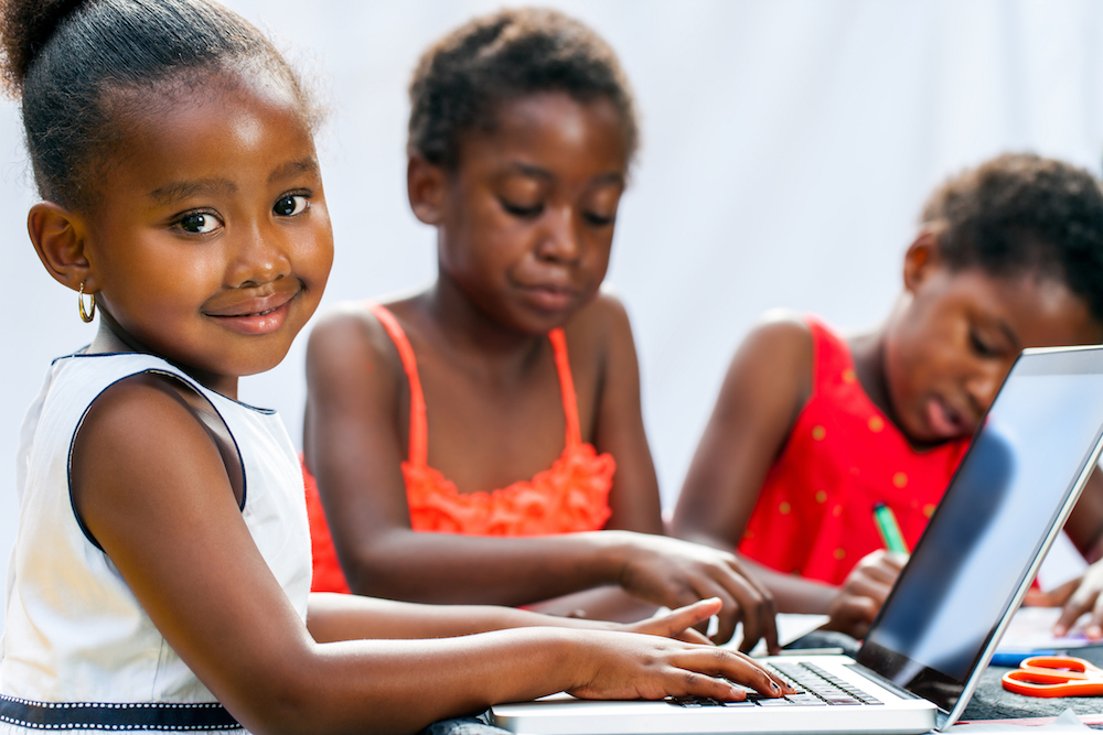 Portrait of cute little african girl coding on computer with friends at desk.Isolated on light background.