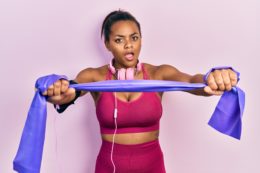 Young african american girl training arm resistance with elastic arm bands in shock face, looking skeptical and sarcastic, surprised with open mouth - sensitive back