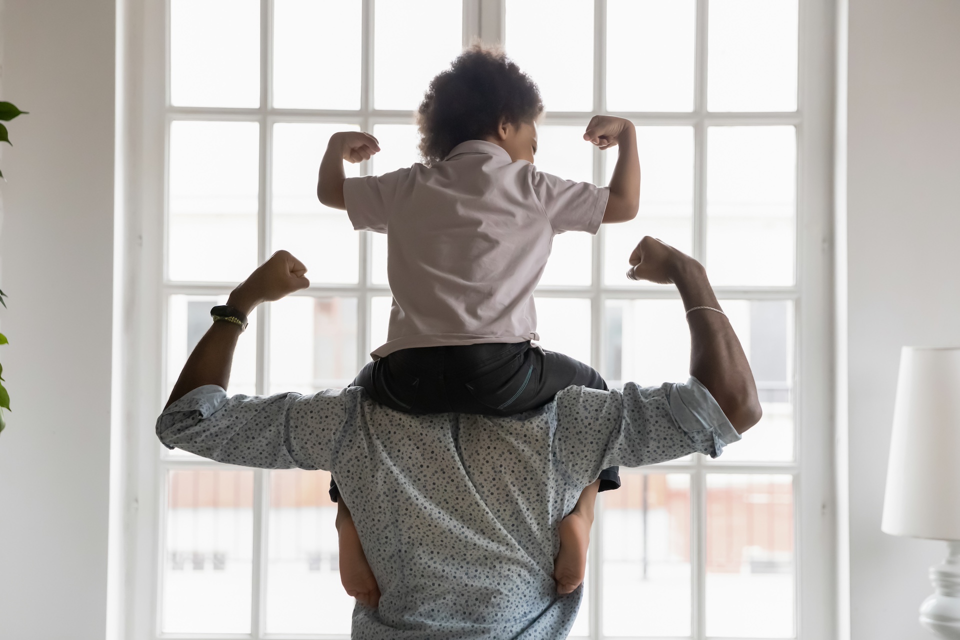 African american dad is protecting a child's life and making the strong symbol with their arms. Father and son in front of a window showing their strength in their arms.