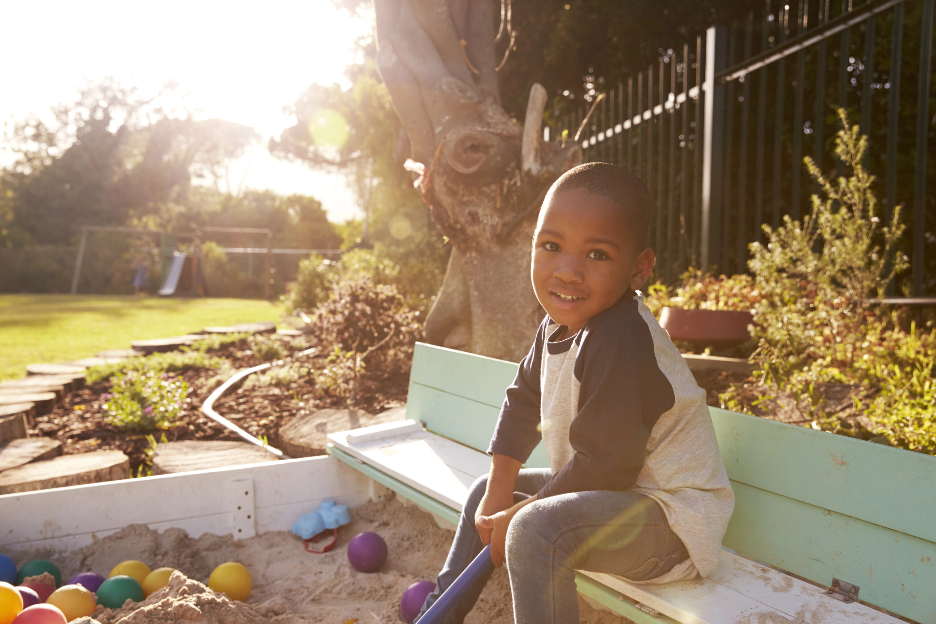 A young boy smiling and sitting in a sandbox filled with colorful balls, in a sunny outdoor setting. The image captures the essence to stimulate young minds through sensory play activities, highlighting the importance of engaging children in hands-on, exploratory play to enhance their cognitive development and senses.