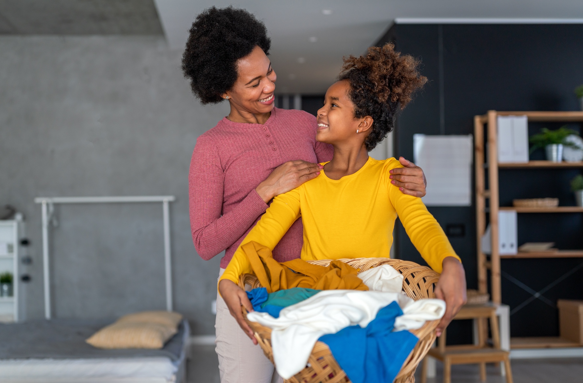 Mother and daughter smiling at each other while holding a basket of laundry, emphasizing teamwork and responsibility to foster independence in kids.