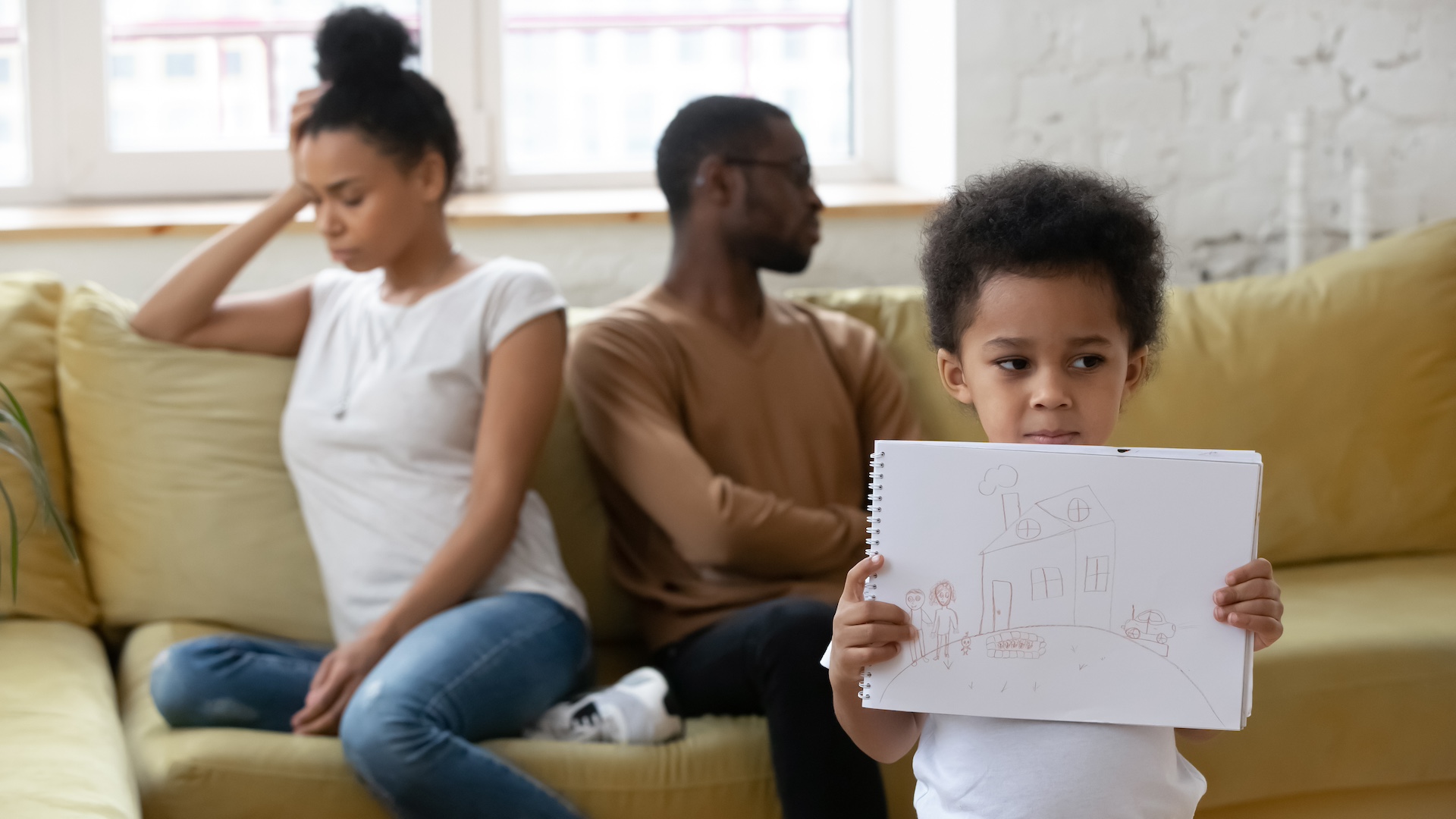 An african american couple is having a sad disagreement in front of their child who is holding a notebook with a happy home for an article on how to keep your kids safe and happy during a divorce