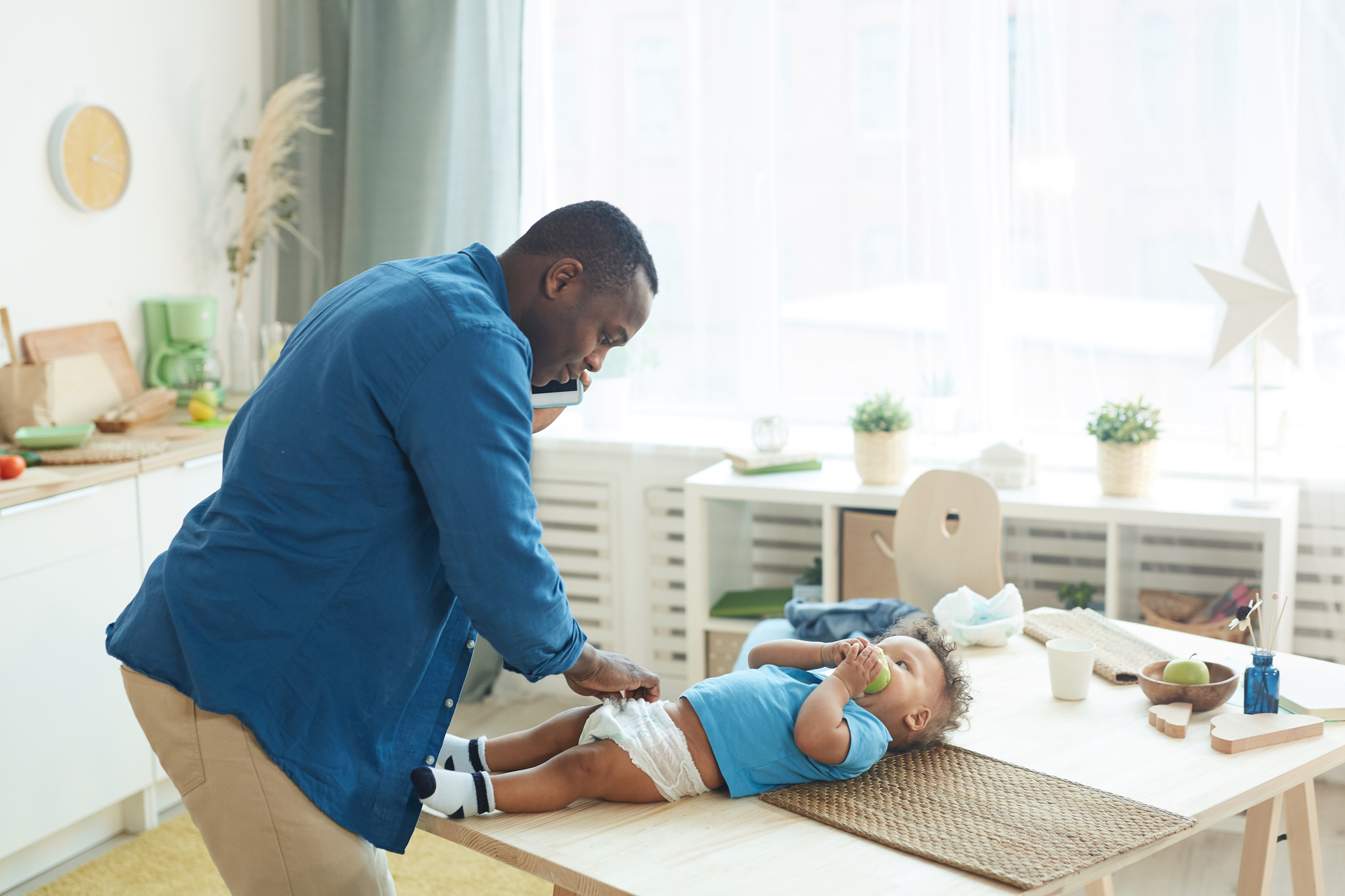 African american dad changes baby diaper for an article on diaper cream. Side view portrait of mature african man calling wife while changing diaper to baby in home interior, copy space