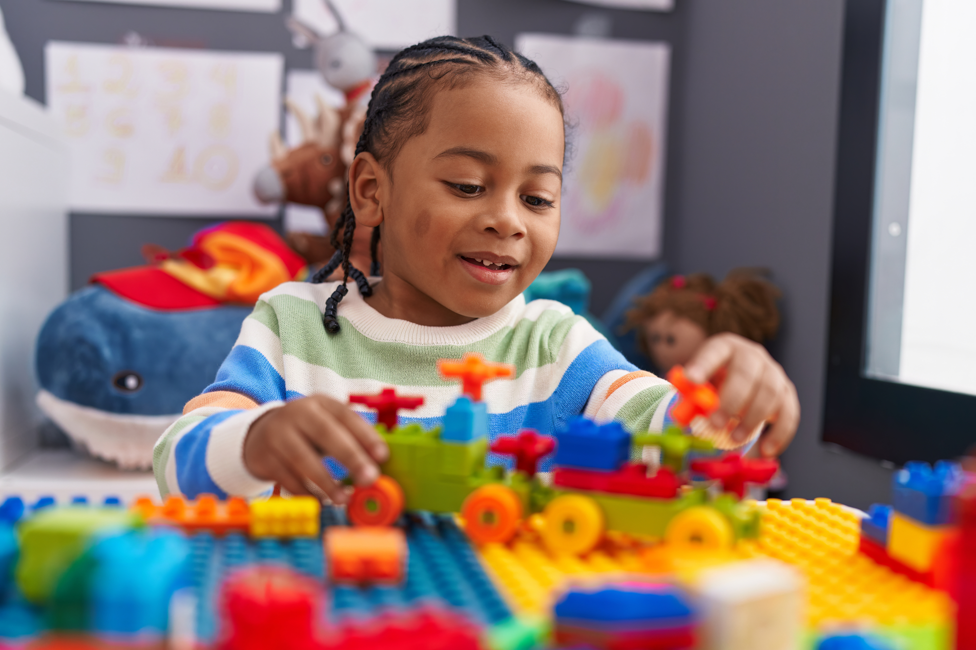 An african american boy with braids is joyfully building with lego bricks at a colorful table. He is wearing a striped sweater and smiling as he assembles his creation, surrounded by toys and a plush whale in the background for an article on lego building tips.