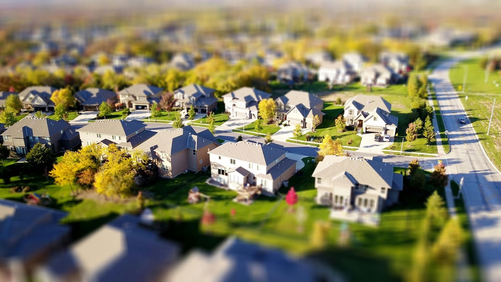 3d aerial neighborhood view of a suburban neighborhood with tree-lined streets, driveways, and homes surrounded by green lawns.