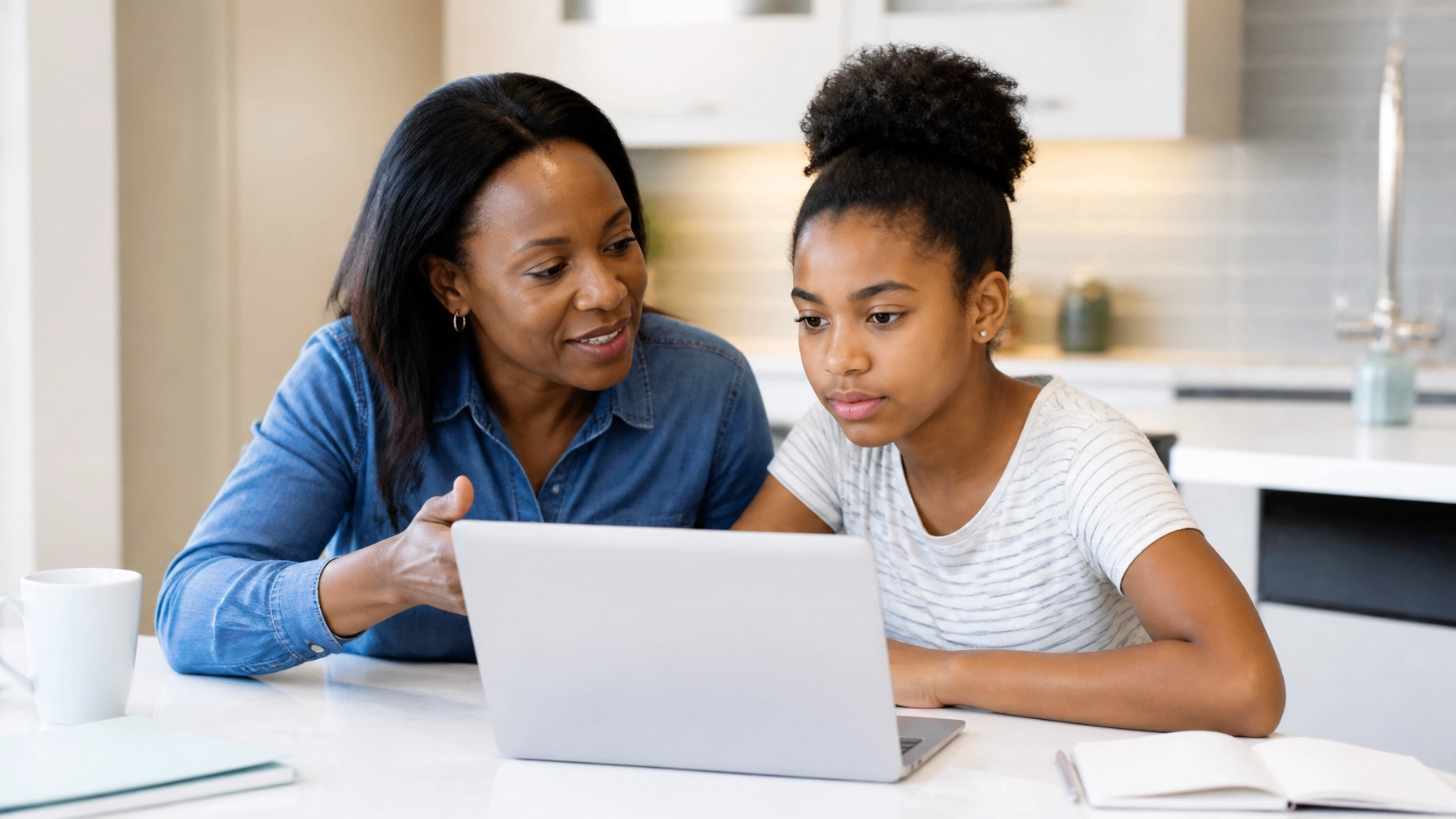 Mother helping daughter navigate laptop at kitchen counter