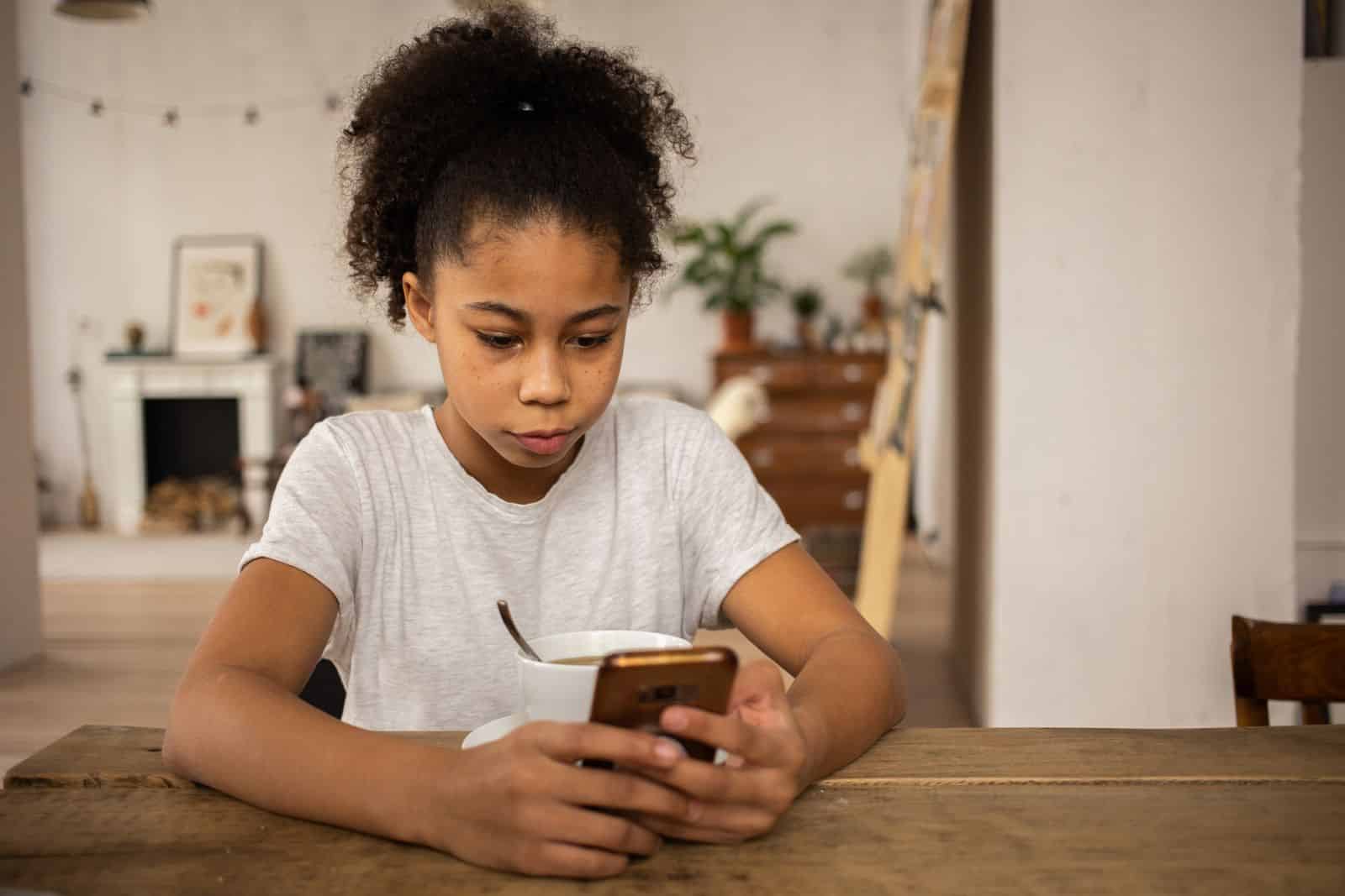 A young girl sits at a table looking at her phone as her family weighs smartphones before middle school.